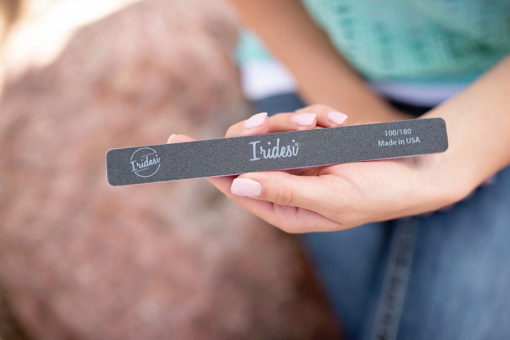 women wearing a turquoise knitted sweater and blue jean, holding an Iridesi printed nail file that has black grit and square end.